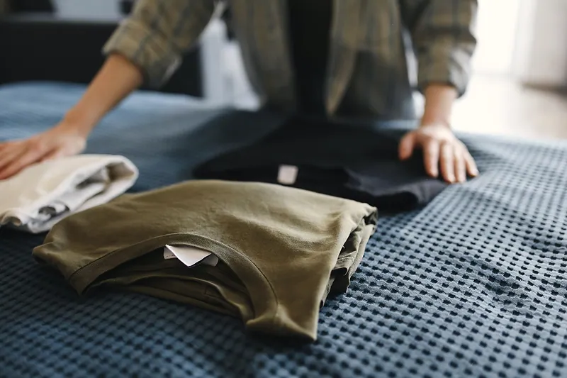 Hands arrange folded T-shirts on a bed.
