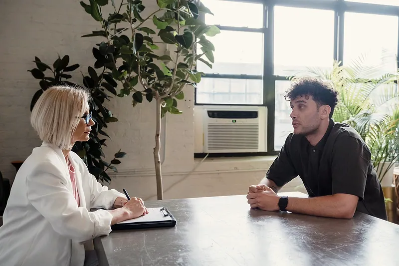 Una mujer con un portapapeles conversa con un hombre en una oficina con plantas.