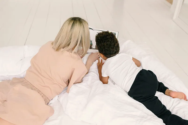 A woman and a boy, seen from behind, read a book together lying on a bed.