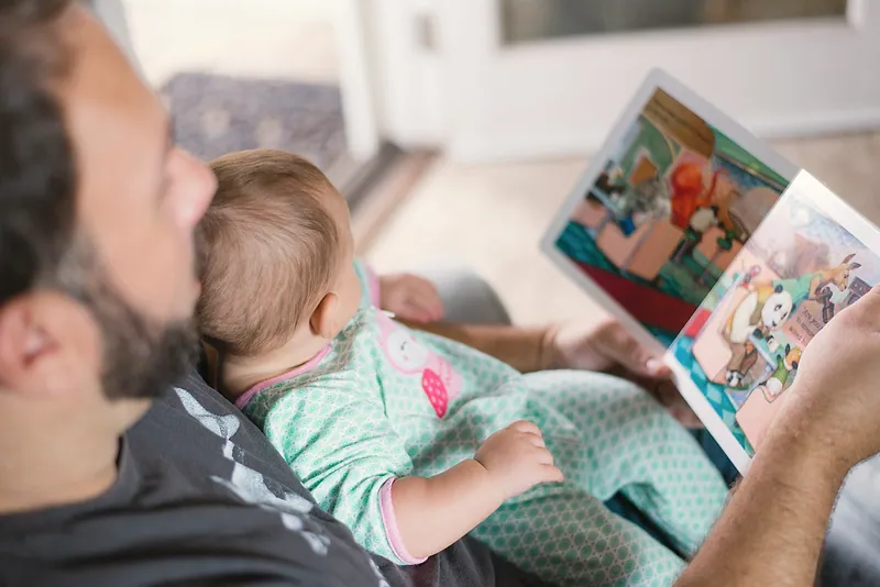 A man reads a picture book to a baby sitting on his lap.