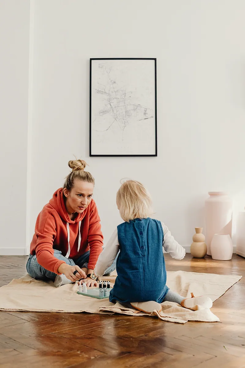 Una mujer juega un juego de mesa en el piso con una niña pequeña que está de espaldas.